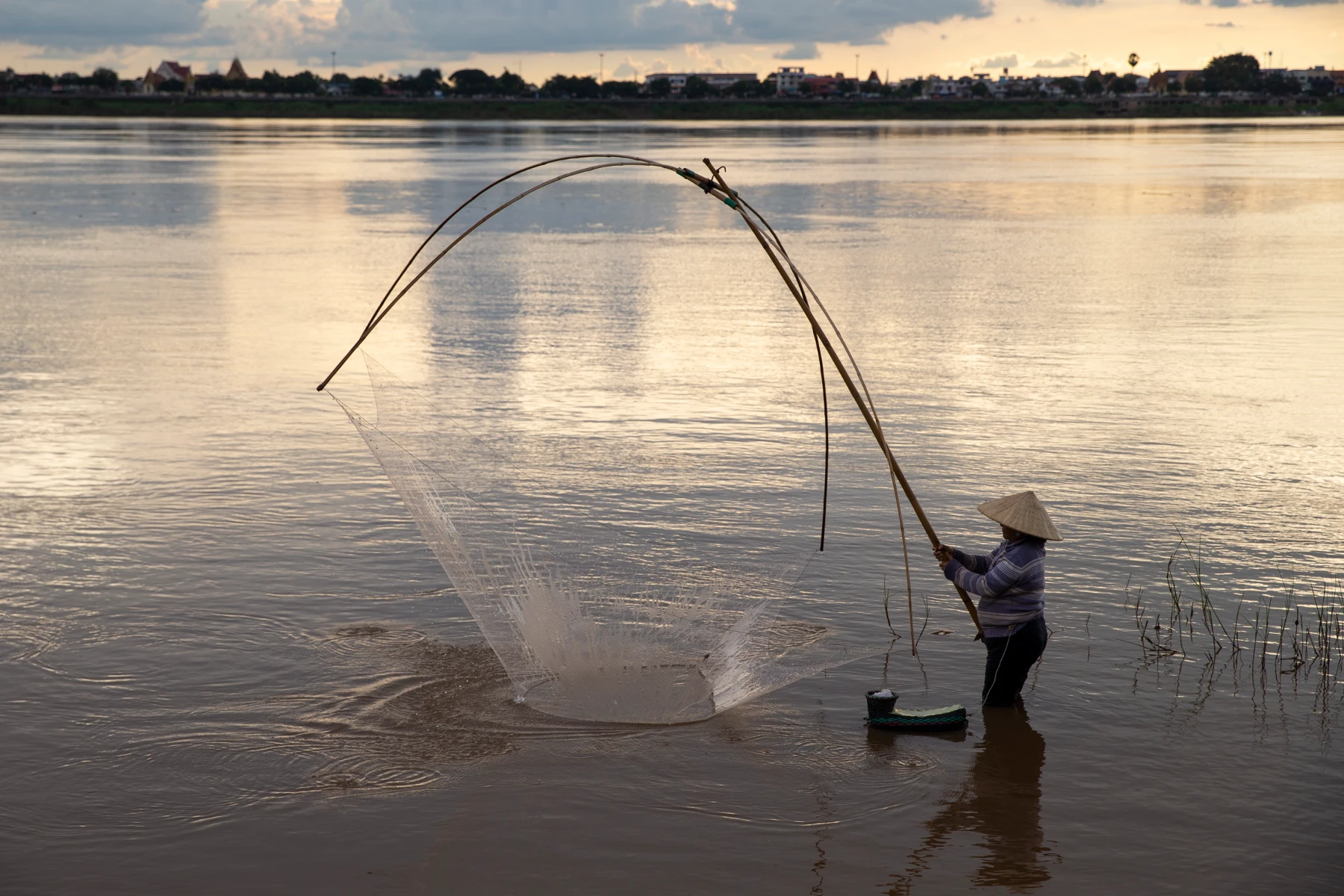 Person wearing a conical hat and striped sweater stands waist-deep in a wide river at sunset, holding long bamboo poles to cast a large circular fishing net that fans out over the water; distant shoreline and soft clouded sky form a low silhouette on the